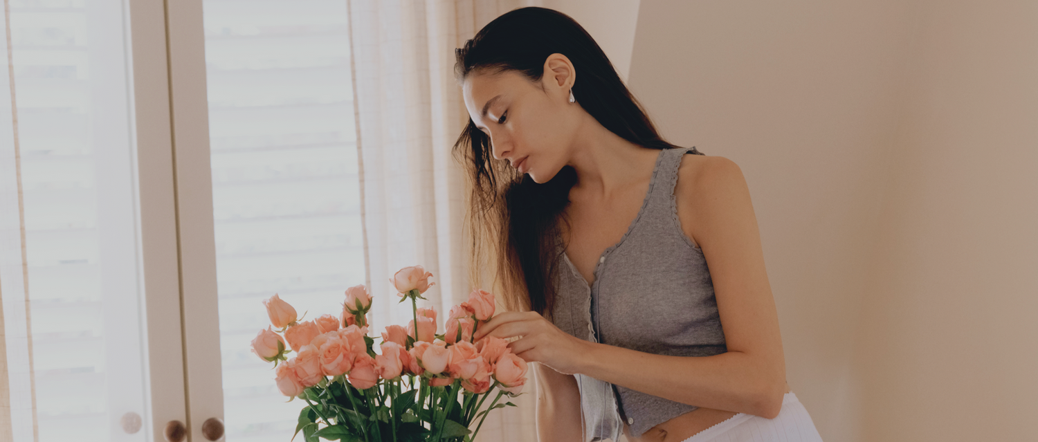 Woman arranging a bouquet of soft pink roses in a naturally lit room, capturing the essence of Gardeny’s rose-inspired cleansing philosophy. The warm, calm atmosphere reflects the Reset & Refresh Mini Duo ritual, melt, hydrate, clean anywhere, symbolizing gentle self-care, plant-based PDRN hydration, and the soothing floral energy behind Gardeny’s Korean cleansing formulas.