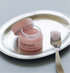 Open jar of Gardeny Rose PDRN Cleansing Balm resting on a polished silver tray with a spatula holding a scoop of its sherbet-like texture. The image highlights the balm’s melt-to-oil formula that dissolves makeup, SPF, and buildup effortlessly while infusing skin with plant-based Rose PDRN for hydration, radiance, and a refreshed, supple, comfortable finish.