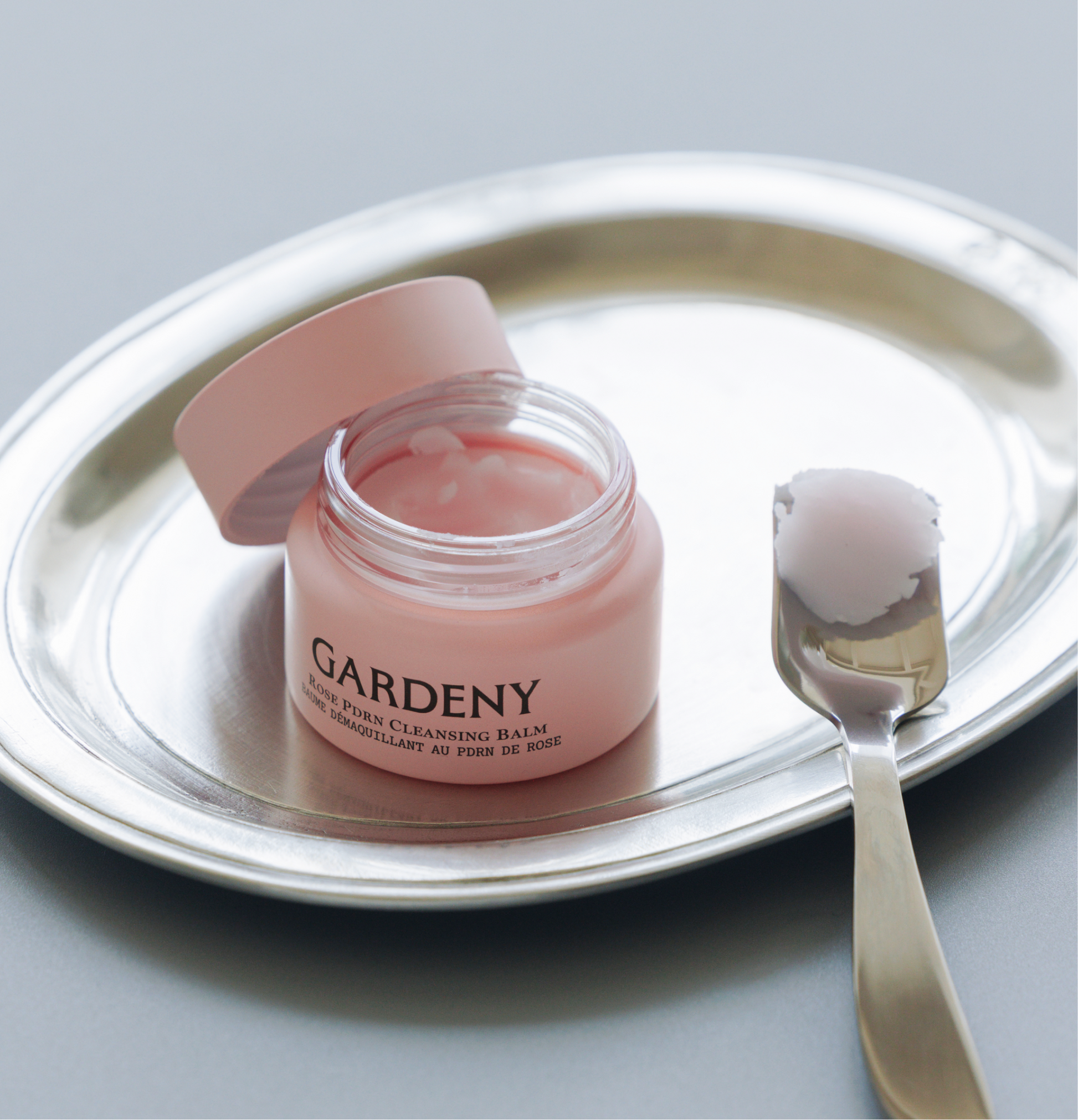 Open jar of Gardeny Rose PDRN Cleansing Balm resting on a polished silver tray with a spatula holding a scoop of its sherbet-like texture. The image highlights the balm’s melt-to-oil formula that dissolves makeup, SPF, and buildup effortlessly while infusing skin with plant-based Rose PDRN for hydration, radiance, and a refreshed, supple, comfortable finish.
