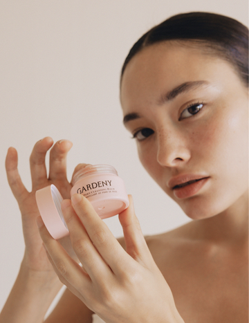 Model opening the Gardeny Rose PDRN Cleansing Balm jar, revealing its silky sorbet-like texture that melts effortlessly into the skin. The image highlights the balm as a travel-friendly, rose-infused cleansing essential designed to dissolve makeup and SPF while delivering deep hydration, radiance, and a soft, supple finish through plant-based Rose PDRN technology.
