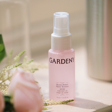 The Gardeny Rose PDRN Mist Toner standing on a softly lit surface next to delicate pink petals and blurred greenery. The translucent pink bottle reflects its hydrating, calming formula enriched with plant-based Rose PDRN, ideal for replenishing moisture on the go, refreshing the complexion, and completing Gardeny’s Essential Mini Trio routine with lightweight, soothing care.