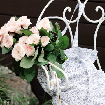 A lush bouquet of pale blush roses with full, delicate petals rests against a vintage white wrought-iron chair, paired with a floral-print Gardeny tote draped over the frame. The muted tones and natural textures evoke Gardeny’s rose-powered skincare essence, reflecting calming nourishment, hydration, and the brand’s garden-inspired beauty philosophy.