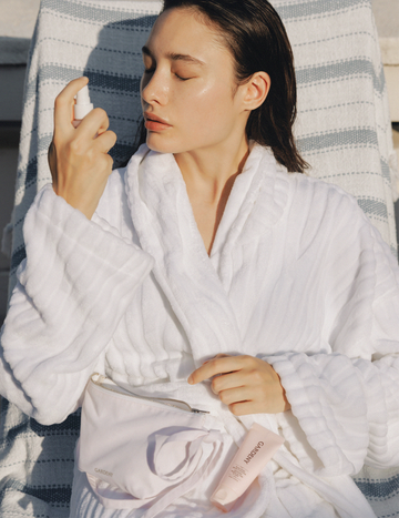 Model in a white robe relaxing outdoors while misting the Gardeny Rose PDRN Mist Toner across the face, with the cleanser and balm resting in a waist pouch. The image emphasizes the trio’s spa-like hydration, calming benefits, and convenient travel-ready format designed to refresh and revive skin with rose-powered moisture wherever you are.