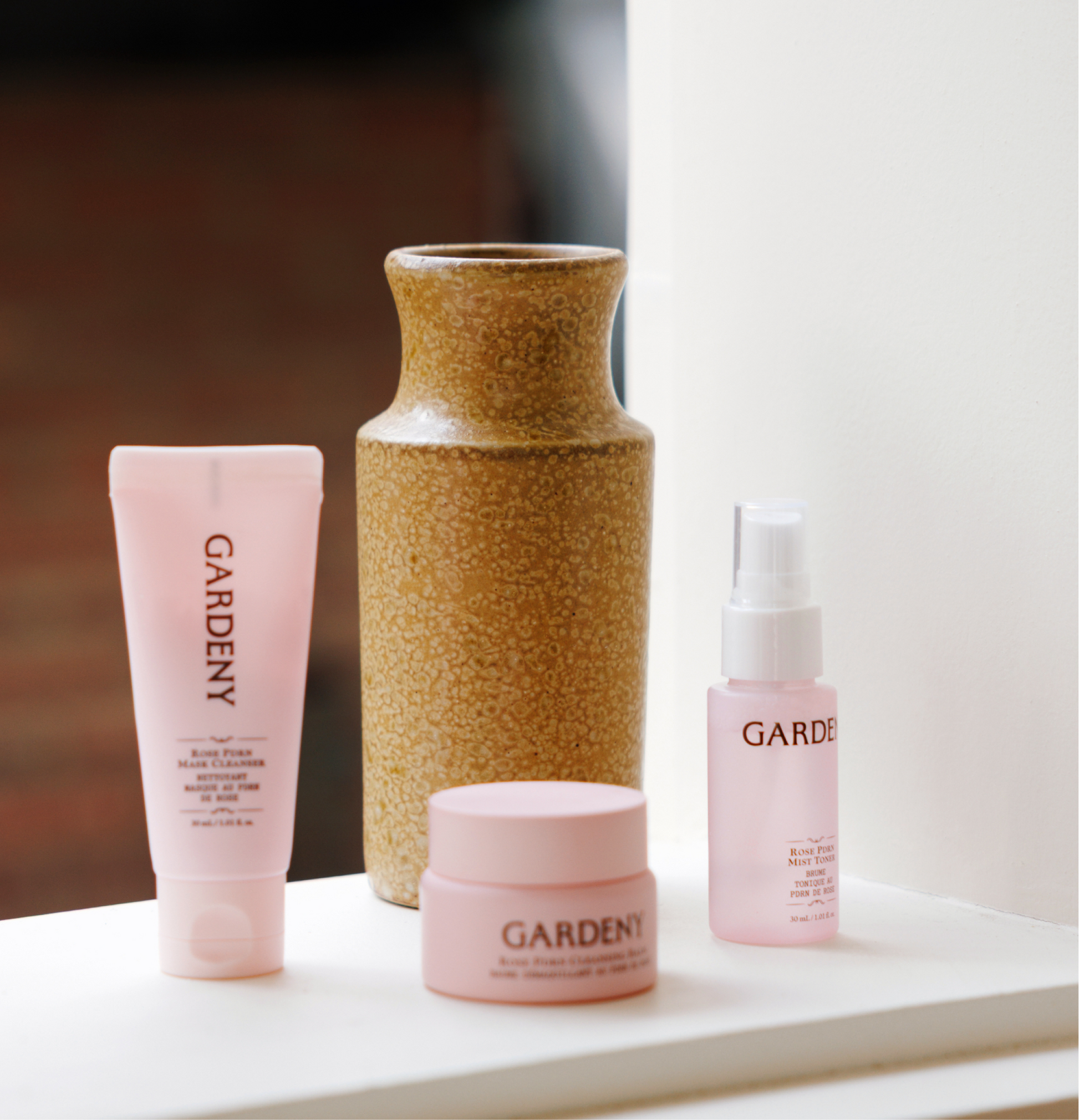 Gardeny Essential Mini Trio arranged beside a ceramic vase on a window ledge in natural light, featuring the mini mask cleanser, cleansing balm, and Rose PDRN mist toner. This setting highlights the trio’s calming, travel-ready design and its ability to restore balance, hydration, and radiance through gentle, rose-infused formulas suitable for everyday routines.