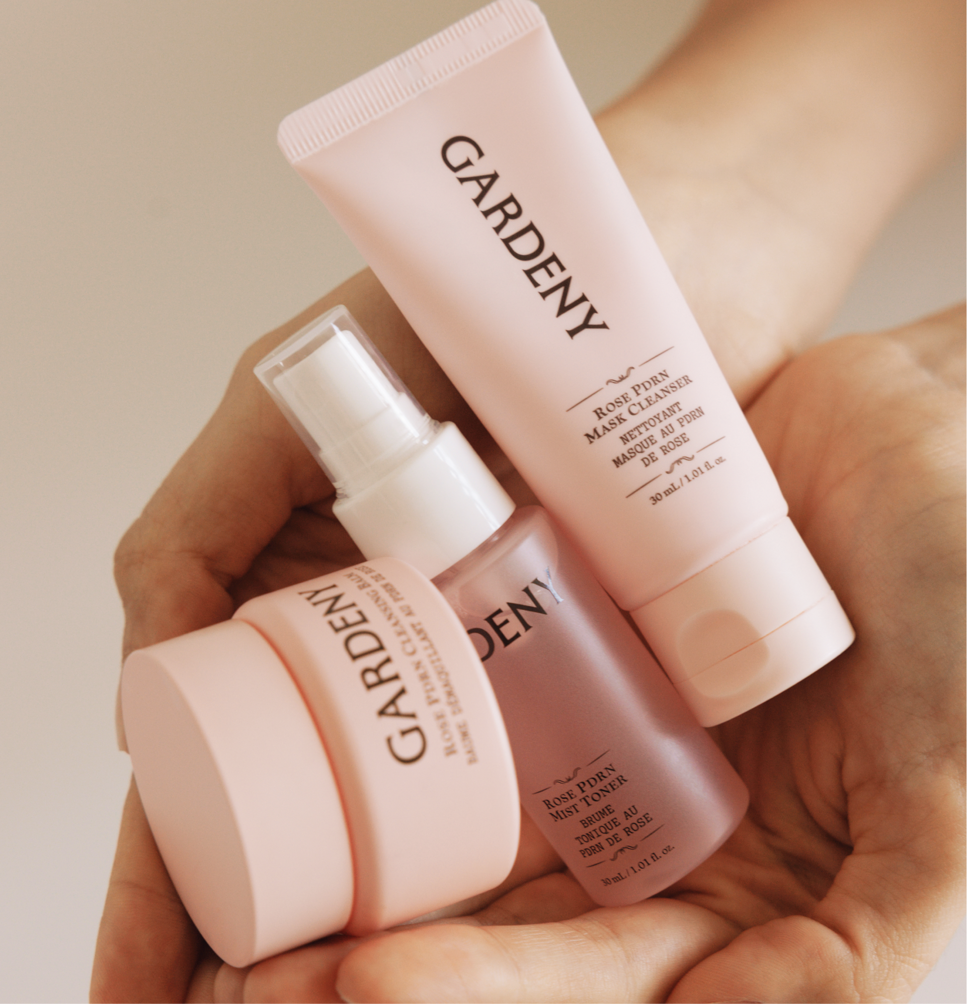 Hands holding the Gardeny Essential Mini Trio, mini cleansing balm, mist toner, and mask cleanser, emphasizing their compact size and soft pink design. The image conveys the convenience of a portable 3-step routine offering gentle cleansing, hydration, and Rose PDRN-powered skin repair, perfect for maintaining a refreshed glow anywhere.