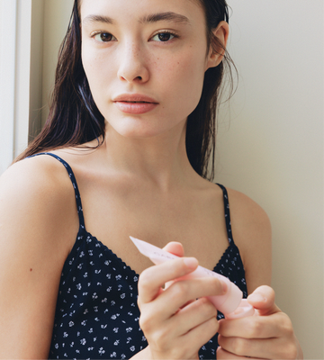 Model holding the Gardeny Rose PDRN Mask Cleanser mini near a bright window, showcasing its lightweight tube and convenient on-the-go size. Her calm expression reflects Gardeny’s easy self-care focus, emphasizing plant-based Rose PDRN formulas designed to cleanse, soothe, and revive skin wherever the day leads as part of the Essential Mini Trio routine.